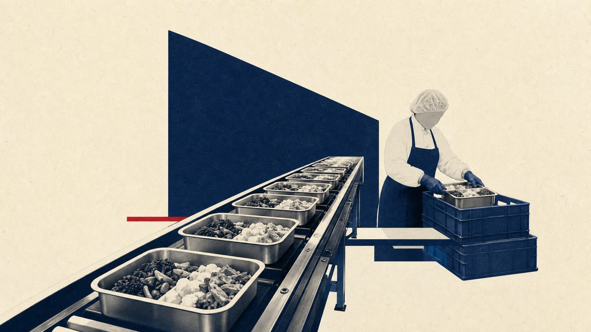 Editorial image of an industrial catering kitchen production line with meal trays on a conveyor belt and stacked meal-packaging crates.